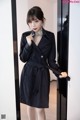 A woman in a black trench coat standing in front of a mirror.