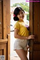 A woman in a yellow shirt and white shorts leaning against a wooden door.