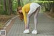 A woman in a yellow sweater and white skirt bending over on a sidewalk.