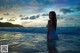 A woman in a blue dress standing in the ocean at sunset.